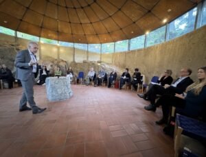 Man in a suit giving a talk to a seated audience in a circular stone room with a wooden domed ceiling and warm lighting.
